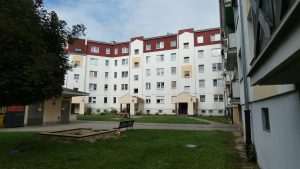 Szeroki widok na duży, pięciopiętrowy blok mieszkalny o symetrycznej architekturze w kształcie podkowy, z białą elewacją i dwuspadowym dachem pokrytym czerwoną blachą. Okna są liczne i w różnych kolorach (białe, żółte). Na parterze w centralnej części widoczne są dwa zadaszone wejścia. Przed budynkiem znajduje się trawnik z ławką i piaskownicą lub małym placem zabaw, otoczony chodnikami.