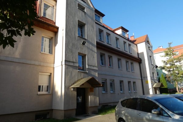 Zbliżenie na fragment jasnego, pięciopiętrowego budynku mieszkalnego, prawdopodobnie kamienicy, z mansardowym dachem w kolorze brązowo-czerwonym. Fasada jest jasna, okna są prostokątne, niektóre z roletami. Widać zadaszone wejście do klatki schodowej oraz fragmenty zaparkowanych samochodów na pierwszym planie. W tle widoczne są inne budynki i drzewa.