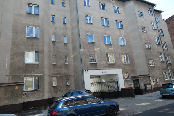 Fasada wielopiętrowego bloku mieszkalnego o szarej, nieco zaniedbanej elewacji. Charakterystyczne są różnorodne okna, niektóre bardzo małe, inne większe, w tym z żaluzjami. Na dole znajduje się duży, biały wjazd do garażu podziemnego lub arkada wejściowa. Przed budynkiem na pochyłej drodze zaparkowane są samochody.