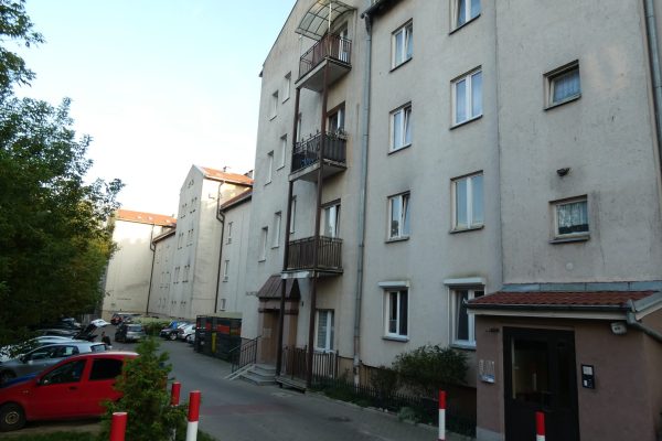 Ujęcie bocznej ściany długiego, wielorodzinnego bloku mieszkalnego w jasnym kolorze. Budynek posiada liczne prostokątne okna i balkony z metalowymi balustradami, niektóre z nich są zadaszone. Na pierwszym planie po lewej stronie znajduje się teren zielony i drzewa, a wzdłuż ściany budynku prowadzi droga z zaparkowanymi samochodami. Widoczne jest również jedno z wejść do budynku.