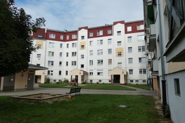 Szeroki widok na duży, pięciopiętrowy blok mieszkalny o symetrycznej architekturze w kształcie podkowy, z białą elewacją i dwuspadowym dachem pokrytym czerwoną blachą. Okna są liczne i w różnych kolorach (białe, żółte). Na parterze w centralnej części widoczne są dwa zadaszone wejścia. Przed budynkiem znajduje się trawnik z ławką i piaskownicą lub małym placem zabaw, otoczony chodnikami.